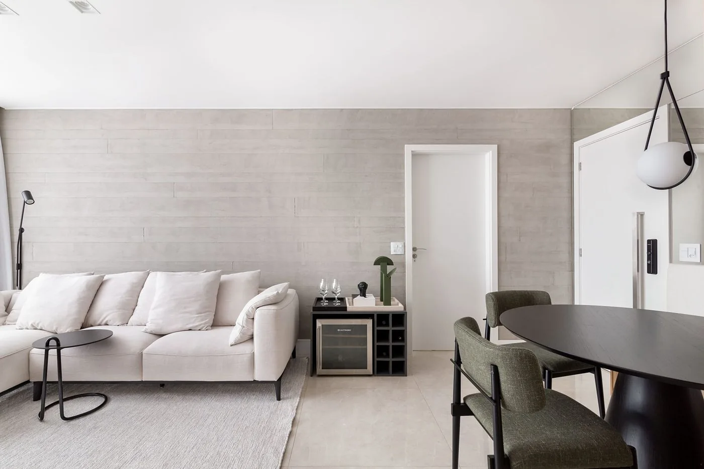 Sala de estar com parede de concreto claro, sofá off-white e mesa de jantar redonda preta com cadeiras verdes