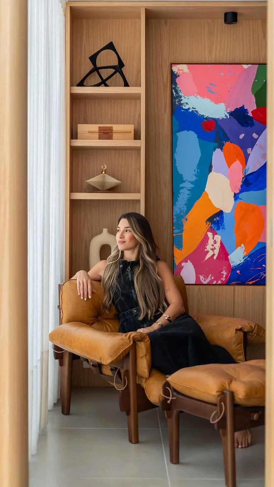 Arquiteta sentada em poltrona de couro caramelo ao lado de uma estante de madeira e quadro colorido
