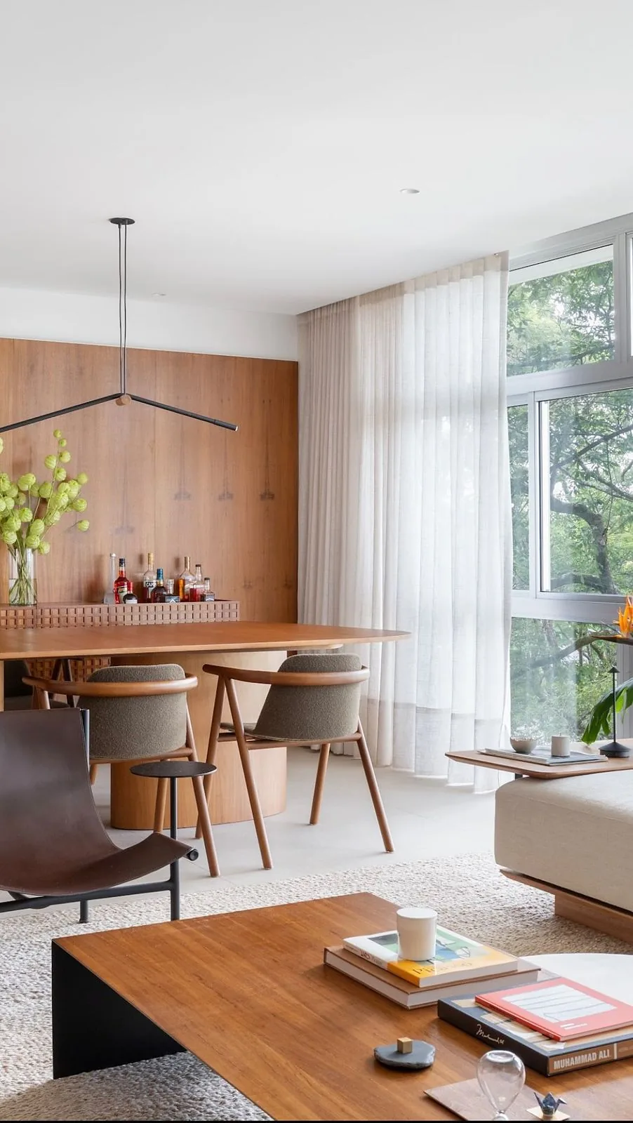 Sala integrada com mesa de jantar em madeira, cortina de linho e janela com vista para árvores
